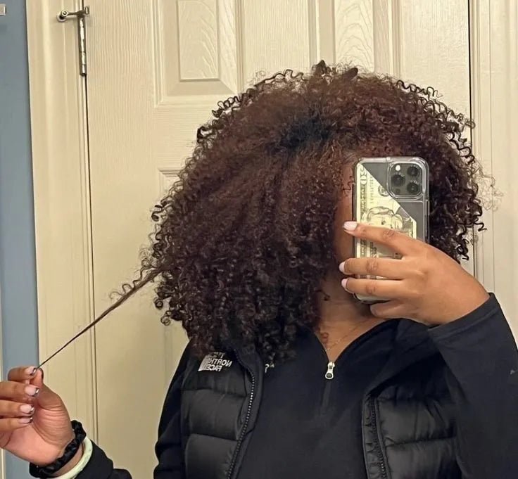 Mirror selfie of a woman with shoulder-length curls, stretching one strand to show its length.